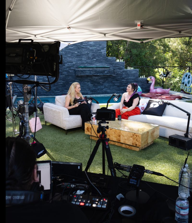 The outdoor podcast set at the house: white couches under a canopy by the pool, camera crew in the foreground